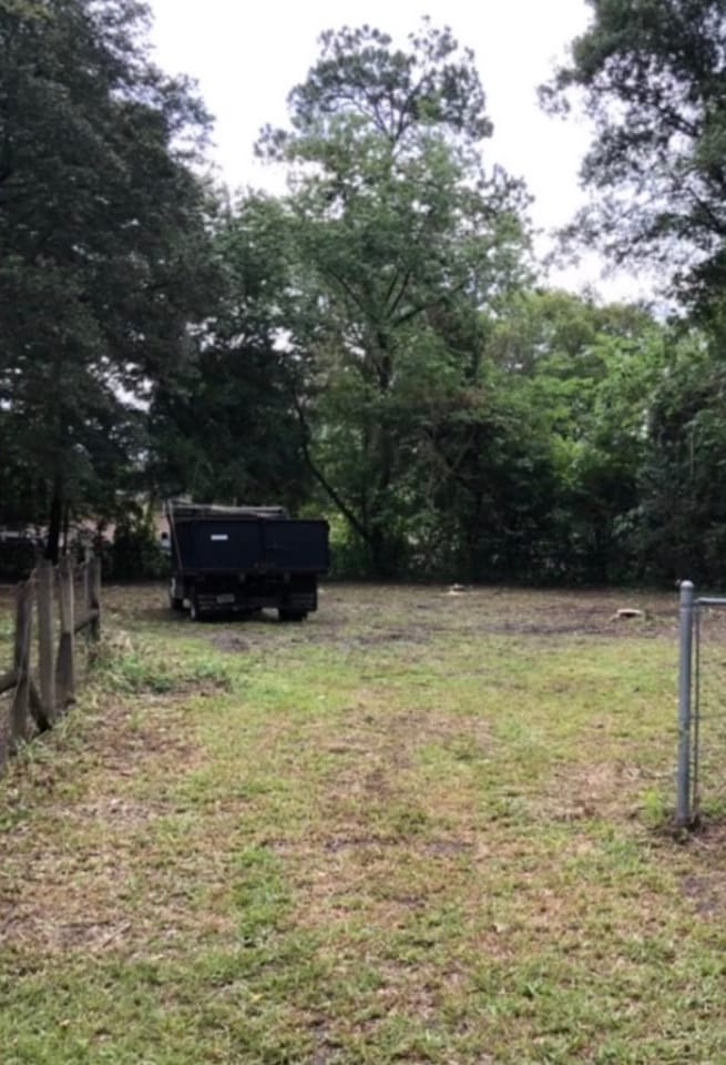 After — the same lot cleared to open grass, with a black dump trailer parked at the tree line.